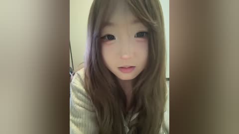 A close-up video of an Asian woman with fair skin and long brown hair, wearing a beige top, looking directly at the camera with a neutral expression.