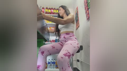 Video of a curvy Latina woman with long dark hair in a white crop top and pink patterned leggings, reaching into a mini fridge, surrounded by colorful snacks and drinks.