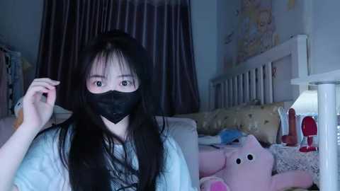 Video of a young Asian woman in a dimly lit bedroom, wearing a black face mask, blue shirt, and long dark hair. Background includes a white bed with a pink plush toy, red dildo, and wall art.