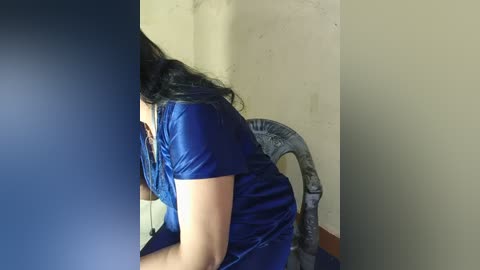 Video of a woman with long black hair, wearing a shiny blue dress, seated on a chair against a beige wall, her back turned.