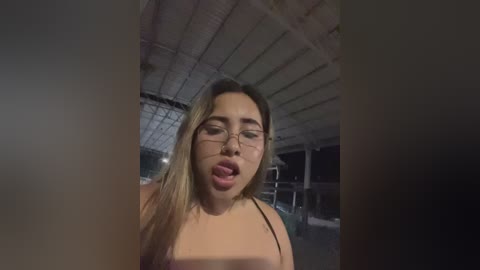 Media: Video of a young woman with long blonde hair and glasses, sticking out her tongue in a playful manner, indoors under dim lighting, wearing a black top.