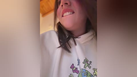 Video of a smiling woman with light skin, dark hair, and wearing a white T-shirt with colorful fish print. Background features a blurred, warm-toned indoor setting.