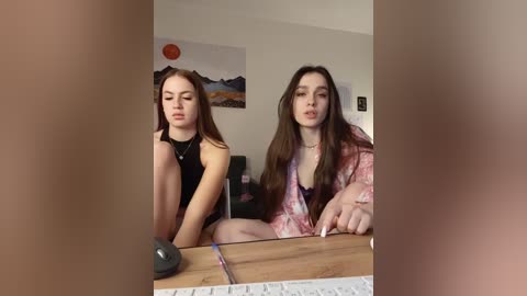 A video captures two young women, one in a black halter top, the other in a pink floral robe, seated at a wooden desk, with a computer monitor and keyboard in view.