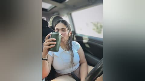 A video of a woman with dark hair, wearing a white top, drinking from a green travel mug while sitting in a car. The background shows a blurred view of the outside.