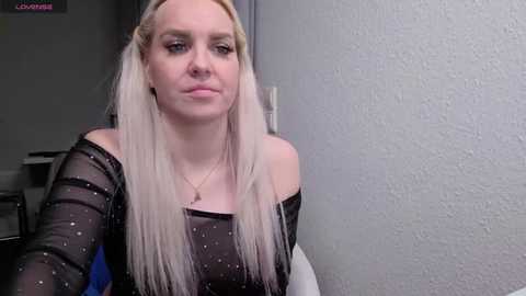 Video of a fair-skinned, blonde woman with long hair, wearing a sheer black top, standing against a textured gray wall.