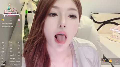 A video of a young Asian woman with fair skin and long brown hair, sticking out her tongue, wearing a white top, taken indoors.