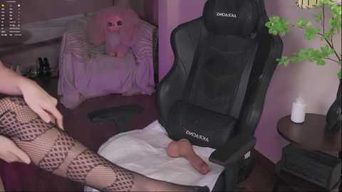 Video of a woman with fair skin, wearing fishnet stockings, massaging a man's erect penis on a white towel, in a dimly lit room with a black gaming chair, pink wall, and potted plant.