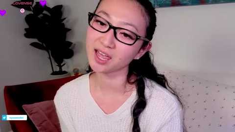 Video of an Asian woman with long black hair, glasses, and a white sweater, in a bedroom with a potted plant and a bed in the background.