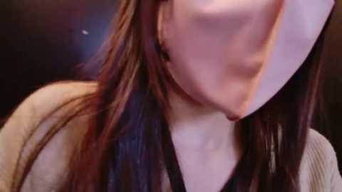 Video of a person with long, straight brown hair, wearing a beige sweater, and a face mask, partially obscuring their face. The background is dark and indistinct.