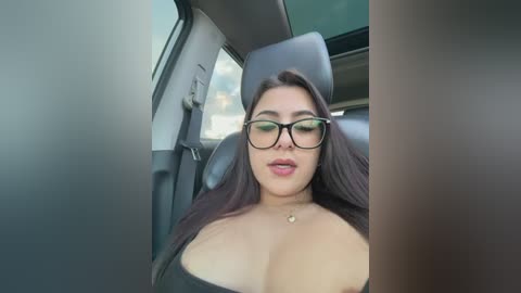 A video of a woman with long dark hair, wearing black-rimmed glasses, a black top, and a gold necklace, sitting in a car.