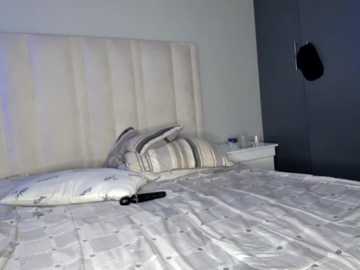 Media: Video of a modern bedroom with a white tufted headboard, striped pillows, white duvet, black sex toy on bed, and a black door with a peephole.