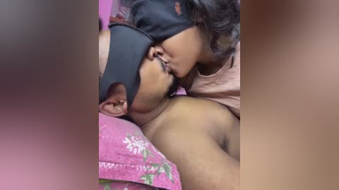 A video of a South Asian man with a black sleep mask on, kissing a woman's cheek while lying on a pink floral pillow, both in intimate, close proximity.
