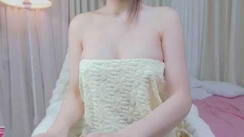 Video of a slender, light-skinned woman with small breasts, wrapped in a white quilted blanket, sitting on a bed with pink sheets. Background includes a white lamp and beige curtains.