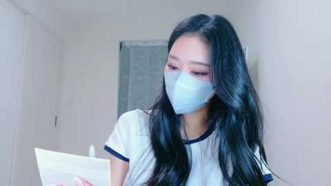 Video of an Asian woman with long black hair, wearing a white T-shirt and blue surgical mask, sitting in a plain white room with a closed door and window.