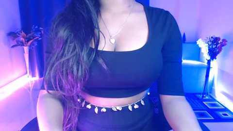 Video of a woman with long, wavy dark hair, wearing a black crop top and high-waisted skirt, sitting in a dimly lit room with purple lighting, blue furniture, and floral arrangements in the background.