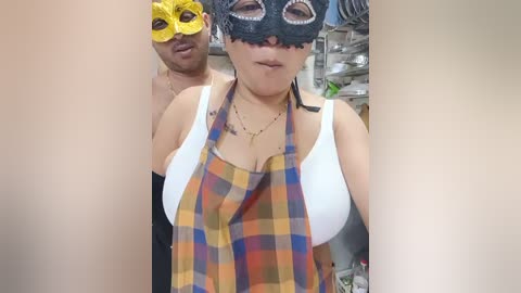 Media: Video of a woman in a white tank top and a yellow plaid apron, wearing a black masquerade mask. A man with a medium build and light skin, wearing a yellow mask, stands behind her. The background features kitchen appliances.