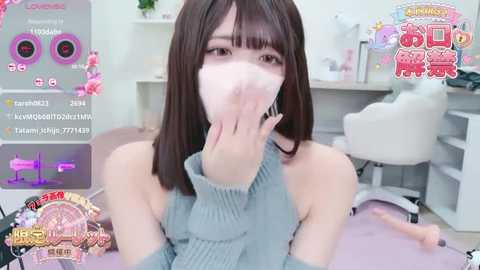 A video of an East Asian woman with straight, shoulder-length dark brown hair, wearing a grey turtleneck sweater and a face mask, sitting indoors in a modern, minimalistic room.