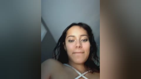 Video of a young woman with medium skin tone, dark curly hair, and a slight smile. She wears a white strappy top. The background is blurred, with a hint of a white wall.