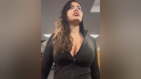 Video of a curvy Latina woman with long, wavy, brown hair in a black, zip-up bodysuit, looking up with a confident expression. Background features a modern, indoor setting with neutral tones and overhead lighting.