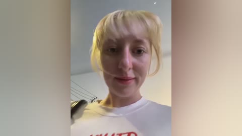 Video of a smiling Caucasian woman with light blonde hair in a white T-shirt, standing in a dimly lit room with a blurred background.