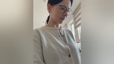 Video of a fair-skinned woman with dark hair, wearing glasses, and a beige cardigan, standing in a bright, modern room with white walls and a window with blinds.