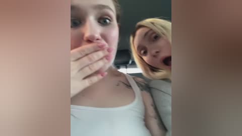 A video of two young women in a car, one with blonde hair and the other with brown hair, both wearing white tank tops, laughing and covering their mouths.