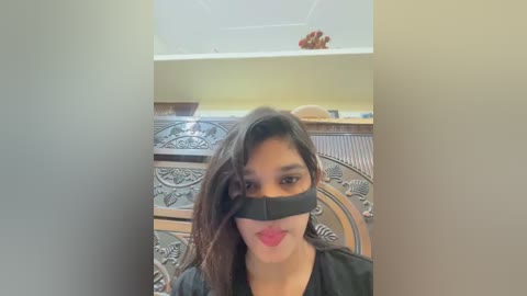 A video of a woman with long, dark hair, wearing a black blindfold and red lipstick, seated in a room with a wooden headboard and a beige ceiling.