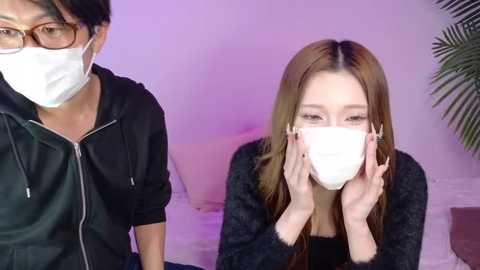 Video of an East Asian woman with long brown hair, wearing a black fur-lined jacket and white mask, sitting on a bed, looking at a man wearing a black hoodie and mask.