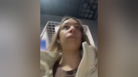 Media: A video of a young woman with blonde hair and fair skin, wearing a beige jacket and a brown top, sitting in a white plastic chair. The background shows a dimly lit room with a metal grid ceiling.