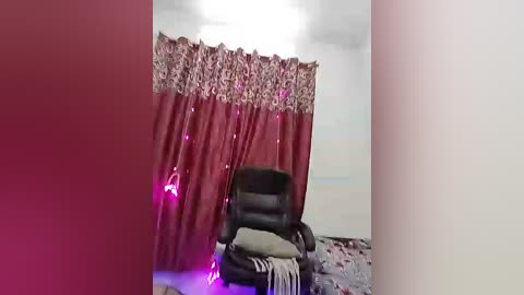 Video of a dimly lit room with maroon curtains adorned with intricate floral patterns, a black leather chair, a grey blanket, and a purple light source casting a soft glow.
