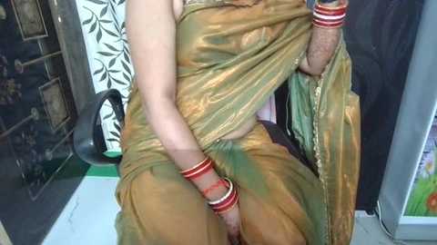 Video of a woman in a green, gold, and red sari, adorned with multiple bangles, sitting on a chair in a room with floral wallpaper and a green floor.