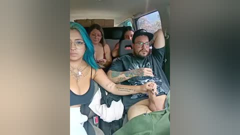 A video captures a man in a car with a woman, both topless, the woman with blue hair and tattoos, the man with a beard and tattoos, in a casual, intimate setting.