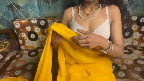 Video of a woman with medium skin tone, wearing a white bra, yellow sari, and multiple pearl necklaces, seated on a patterned couch with retro decor.