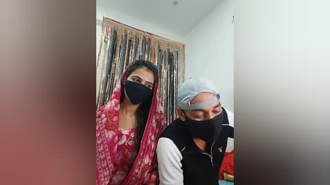 Video of two women, one in a pink floral dress, the other in a black hoodie, standing in a hallway with a beige curtain and colorful fabric hanging in the background.