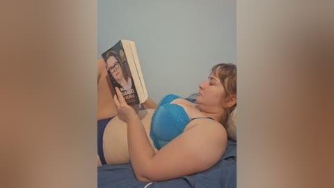 Media: Video of a plus-sized woman with light skin, brown hair, wearing a blue bra, reading a book on a blue bedspread in a minimalistic room.