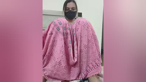 Media: A video of a woman with dark hair and glasses wearing a black face mask and a pink, intricately patterned, lacy blanket covering her breasts. The background shows a plain, white-walled room.