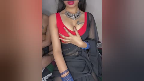 A video of an Indian woman with medium skin tone, wearing a red bra and black sari, with silver jewelry and a blindfold. She is holding her breast, revealing a tattoo.