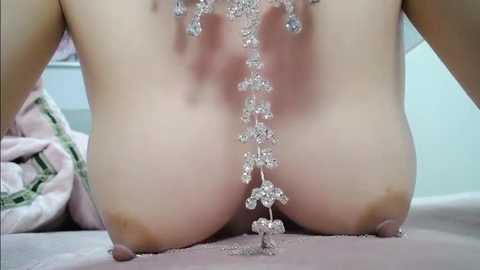 A close-up video of a nude woman with light skin, wearing a silver chain necklace with dangling charms, positioned on her back on a pink bedspread.
