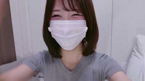 A video of an East Asian woman with straight, shoulder-length brown hair, wearing a white surgical mask, gray t-shirt, and a neutral expression. The background features a plain white wall and part of a bed.