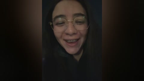 A dimly lit video shows a young woman with long black hair, wearing round glasses and braces, smiling broadly with closed eyes. She has fair skin and is dressed in a dark outfit.