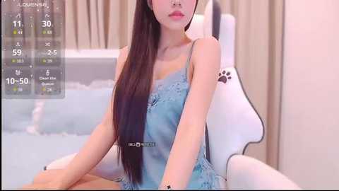 Video of an Asian woman with long black hair, wearing a blue lace camisole, sitting on a white gaming chair in a minimalist room with beige curtains.