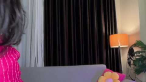 Video of a woman with shoulder-length, wavy gray hair in a pink, textured blouse, standing in a room with black curtains, a white lampshade, and a potted plant.