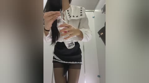 Video of a slender Asian woman in a maid outfit with a black mini skirt, white blouse, and sheer black stockings. She's pouring water into a clear bottle in a narrow hallway.