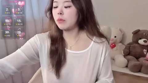 Video of an Asian woman with long brown hair, wearing a white top, sitting on a bed, surrounded by plush toys, including a bear.