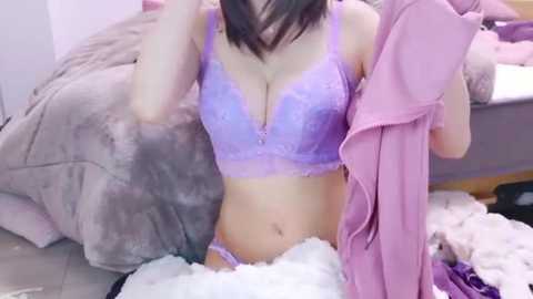 Video of an East Asian woman with long dark hair, kneeling on a white faux fur rug in a messy bedroom. She wears a lavender lace bra and panties, holding a pink shirt.