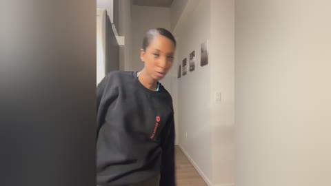 Video of a young Black woman with a slender build and short hair, wearing a black sweatshirt with red text, standing in a minimalist hallway with white walls and framed photos.