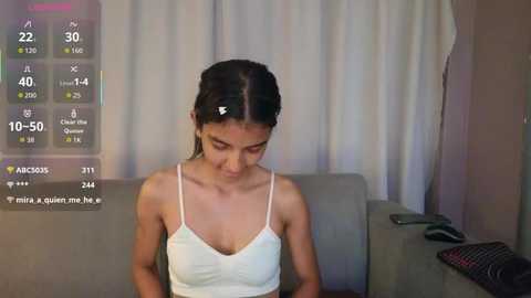 Video of a young Asian woman with straight black hair tied back, wearing a white tank top, sitting on a grey couch in a dimly lit room. A digital overlay displays heart rate, steps, and calories burned.