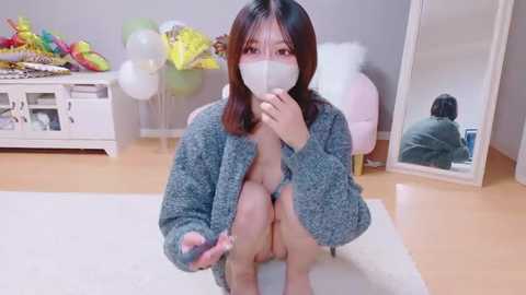 Video of an Asian woman with shoulder-length black hair, wearing a gray cardigan, white mask, and no pants, squatting on a white rug in a bright, modern room with a white chair and colorful balloons.
