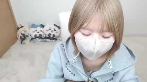 Video of a young Asian girl with short blonde hair, wearing a white face mask, light blue hoodie, and sitting on a beige carpet. Background features stuffed panda toys and a wooden door.