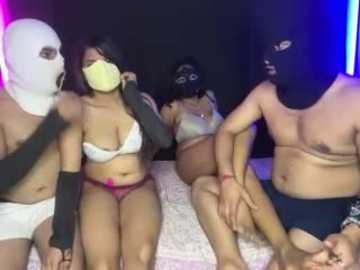 Video of five individuals in BDSM attire: four wearing masks, one with yellow tape over mouth. They sit on a bed, partially clothed, with a black and blue-lit background.